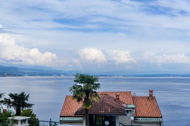 Opatija, Hırvatistan - 27 Mayıs 2024: Önünde terracotta fayans çatısı ve palmiye ağaçları olan kıyı evi, uzaktaki bulutlu sakin bir denize bakıyor. Sahne, deniz kenarındaki bir yerin güzelliğini yakalar..