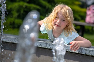 Sarı saçlı genç bir kız çeşmenin üzerinden eğilip eliyle suya uzanıyor. Açık mavi bir gömlek giyiyor ve parlak güneş ışığı sudan yansıyor ve kızların saçı....