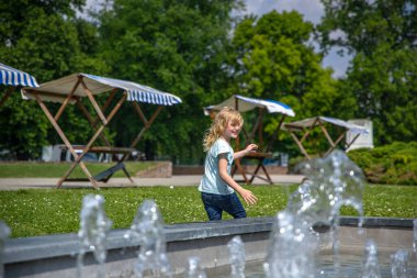 Kot pantolonlu ve açık mavi gömlekli genç bir kız şehir parkındaki bir çeşmenin önünden geçiyor, gülüşü gün ortası güneşinin altında parlıyor..