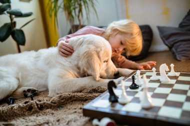 Köpeği kafasını tahtaya dayayıp dikkatle izlerken küçük bir kız yerde satranç oynuyor..