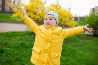 Genç bir çocuk çimenli bir parkta kollarını açmış duruyor. Sarı yağmurluk ve örgü şapka giyiyor..