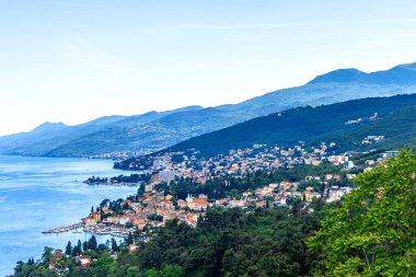 Hırvatistan 'ın Opatija kıyı şeridinin manzarası, açık mavi bir gökyüzü, Adriyatik Denizi, yeşil tepeler ve ağaçların arasına yuva yapmış bir kasaba.