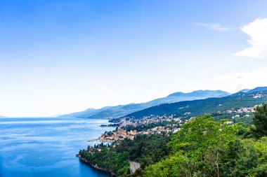 Opatija kıyı şeridinin panoramik manzarası, yeşil tepeler ve ışıl ışıl mavi Adriyatik Denizi arasına kurulmuş kasabayı gösteriyor. Açık bir gökyüzü ve yemyeşil bitki örtüsü resim gibi bir ortam yaratır..