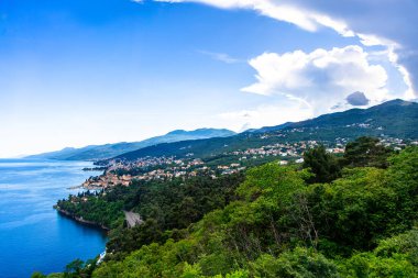 Opatija kıyı şeridi manzaralı, yemyeşil bitki örtüsü, mavi su ve tepelerde yuva yapmış küçük bir kasaba..