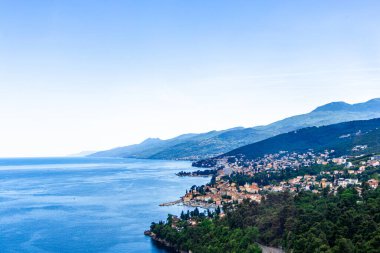 Hırvatistan 'ın Opatija kıyı şeridinin manzarası, açık mavi bir gökyüzü, uzak dağlar ve hareketli bir kıyı kasabası.