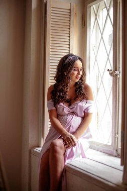 Huzurlu ve düşünceli, uzun kıvırcık saçlı hamile bir kadın pencere pervazında oturuyor, elleri pembe bir elbiseyle karnını sarıyor, küçük kızının gelmesini beklerken, sessiz bir hayalle kayboluyor. Hamilelik kavramı, içgözlem ve antika