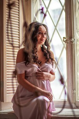 Sessiz bir yansıma anında, hamile bir kadın pencere pervazında oturur, uzun kıvırcık saçları yüzünü yumuşakça çerçeveler, elleri yavaşça pembe bir elbisenin içindeki bebek şişliği üzerinde dinlenir. Annelik düşüncesi, beklenti ve sükunet kavramı.