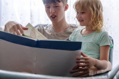 Genç bir adam bir sandalyede oturuyor, kucağında oturan küçük bir kıza kitap okuyor. Kız gülümseyerek kitaba bakıyor ve adam da sayfaları parmağıyla işaret ediyor..