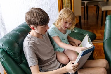 Kahverengi saçlı genç bir çocuk yeşil deri bir kanepede oturuyor ve küçük kız kardeşine kitap okuyor. Kız kardeşi onun kucağında oturuyor ve o okurken sayfaları işaret ediyor. Uzun sarı saçları var ve açık mavi bir tişört giyiyor..