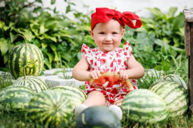 Kırmızı beyaz karpuz desenli elbise giyen genç bir kız yeşil bir yaz bahçesinde karpuz tarlasının arasında oturuyor. O, büyük bir yay ile kırmızı bir saç bandı takıyor ve ağzına bir dilim karpuz tutuyor..