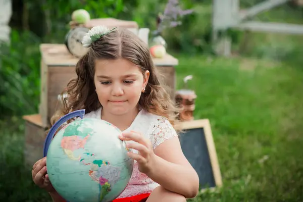 stock image A young girl sits on a wooden crate in a grassy area, holding a globe in her hands and looking at it intently.. Back to School concept