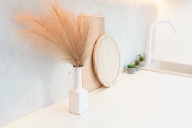 Beyaz bir vazo ve kurutulmuş otlar tezgahın üzerinde duruyor. Tezgahta tahta kesme tahtası ve yuvarlak servis tepsisi de var..