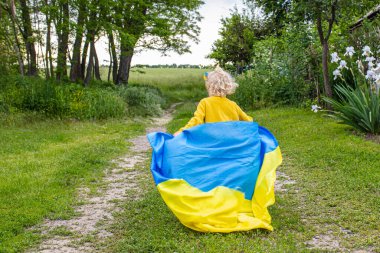 Sarı tişörtlü küçük bir çocuk, arkasında Ukrayna bayrağı taşıyan bir toprak yolda koşuyor..