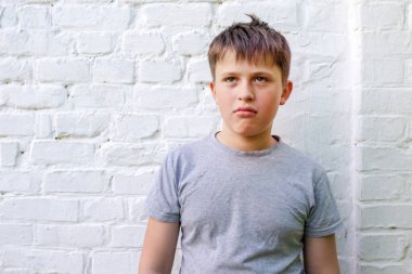 Kahverengi saçlı genç bir çocuk beyaz bir tuğla duvarın önünde duruyor. Gri bir tişört giyiyor ve yüzünde ciddi bir ifade var..