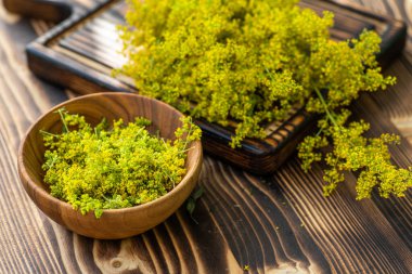 Ahşap bir kasede, sarı bir bayanın karyola çiçekleriyle dolu bir tahta masa üzerinde oturuyor. Daha fazla çiçekli tahta kesme tahtası kasenin yanında yatıyor..