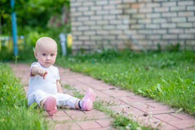 Küçük bir çocuk yeşil çimenli bir arka bahçedeki kırmızı tuğla bir patikada oturuyor. Çocuk beyaz bir gömlek ve pembe ayakkabılar giyiyor ve tek eliyle işaret ediyor..