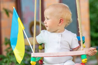 Bir çocuk tahta bir salıncakta oturur, bir elinde küçük bir Ukrayna bayrağı taşır..