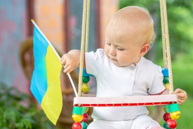 Bir bebek dışarıda salıncakta oturuyor, elinde Ukrayna bayrağı var..