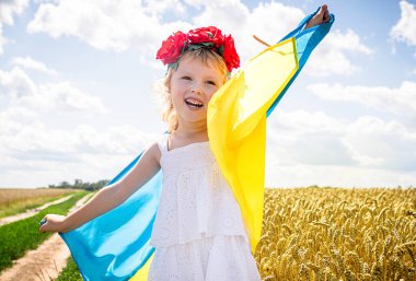 Çiçekli taç giyen neşeli bir çocuk, altın buğday tarlasında dururken Ukrayna 'nın mavi ve sarı ulusal bayraklarını tutuyor. Çocuk güzel kırsalda mutluluk ve gurur ifade ederken güneş ışıl ışıl parlıyor..