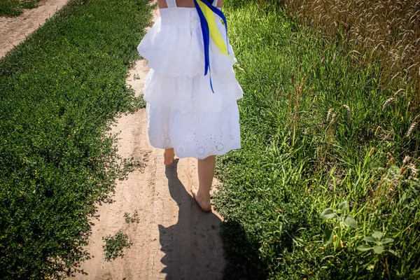 stock image A young person walks barefoot along a dirt pathway, adorned in a flowing white dress. Bright blue and yellow ribbons accentuate the outfit, symbolizing national pride  