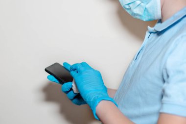Sterilizatörlü akıllı telefonun çocuk dezenfekte edilmesi. Eldivenli eller alkolle ıslanmış beyaz bir bez, evde dezenfektan cep telefonu. Coronavirüs salgını, covid-19 konsepti.