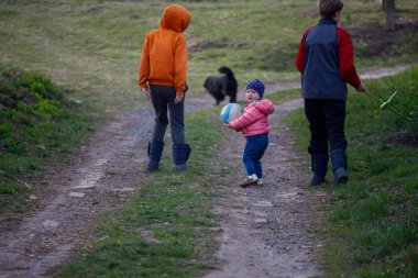 Bir grup çocuk neşeyle bir top taşıyan bir çocukla manzaralı, çimenli bir yolda yürüyor. Oyuncu bir köpek onları takip ediyor, öğleden sonraki maceralarına neşe katıyor..