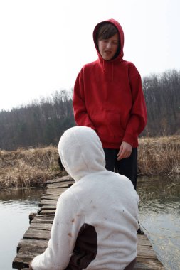 İki çocuk sakin suların üzerindeki, kırsal ahşap bir köprüde duruyorlar, etrafları uzun otlar ve ağaçlarla çevrili, kırsalda sakin bir anın tadını çıkarıyorlar..