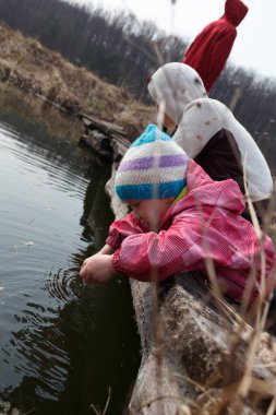 Renkli çizgili şapkalı meraklı bir çocuk göletin kenarında oynuyor, suya uzanıyor, kırsal kesimde serin bir günde iki figür uzaktan izliyor..