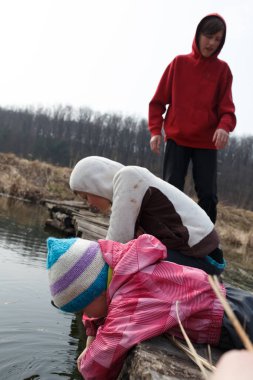 Three children lean over a wooden log at the pond's edge, with one reaching into the water while others watch. They are dressed warmly for the chilly weather. clipart