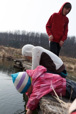 Üç çocuk, bir genç gözlemci ile bir göleti keşfetmek için tahta bir tahtanın üzerine yaslanıyor. Erken bahar manzarası tomurcuklanan doğayı ve yumuşak güneş ışığını gözler önüne serer..