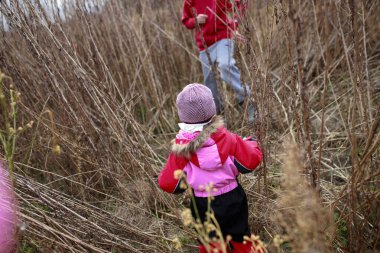 Pembe ceketli ve bereli bir çocuk uzun otların arasında dolaşırken bir yetişkin yanında duruyor ve soğuk sonbahar havasının tadını çıkarıyor..