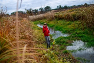  Kırmızı ceketli genç çocuklar, bulutlu bir günde, uzun otlar ve yeşilliklerle çevrili küçük bir derenin yanında, doğanın ve açık havanın tadını çıkarıyorlar..
