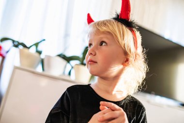 Siyah elbiseli bir çocuk ve kırmızı şeytan boynuzları dikkatle duruyor, neşeli bir toplantı sırasında yan tarafa bakıyor.