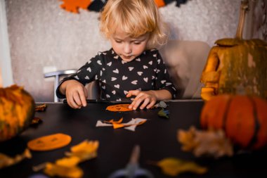 Genç bir çocuk renkli kesikler, balkabakları ve el işi malzemeleriyle dolu bir masada Cadılar Bayramı süslemeleri yapmaya odaklanır. Atmosfer şen şakrak, tatil için mükemmel..