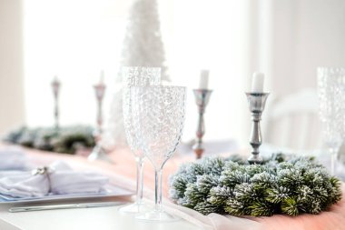 Güzelce düzenlenmiş bir tatil masasında kristal bardaklar, gümüş şamdanlar ve buzlu yeşillik manzarası yer alıyor. Aydınlık oda kutlamanın atmosferini arttırır..