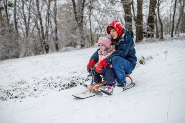 İki çocuk karla kaplı bir tepeden kayıyor, etrafları uzun ağaçlarla çevrili. Sıcak kışlık giysiler içinde serin bir öğleden sonra neşeyle gülüyorlar..