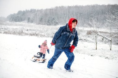 Bir baba, genç kızını karlı bir yolda kızağa çeker. Kış sahnesinde düşen kar taneleri ve arka planda sakin bir orman, neşeli bir atmosfer yaratıyor..