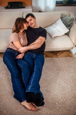 Bir çift, yumuşak bir halıda birlikte rahatlar, sıcak aydınlatılmış bir oturma odasında birlikte vakit geçirirler. Gülüşleri ve nazik dokunuşları Sevgililer Günü için mükemmel bir ortam yaratıyor..