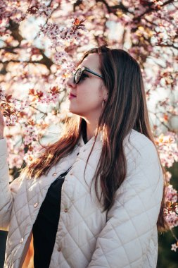 Parktaki kiraz ağaçlarının altında zarif bir şekilde duran bir kadın. Güneş gözlüklerini sergiliyor. Örtülü bir ceket.. 