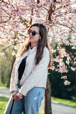 Parktaki kiraz ağaçlarının altında zarif bir şekilde duran bir kadın. Güneş gözlüklerini sergiliyor. Örtülü bir ceket.. 