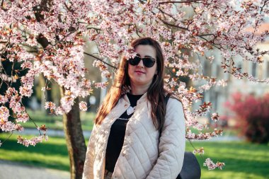 Parktaki kiraz ağaçlarının altında zarif bir şekilde duran bir kadın. Güneş gözlüklerini sergiliyor. Örtülü bir ceket.. 