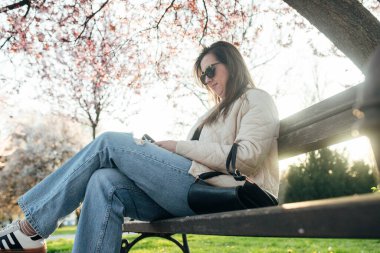 Parktaki bankta rahat bir şekilde oturan, güneşli bir günde telefonunu kontrol eden bir kadın. Etrafında kiraz çiçekleri açarak baharın sakin atmosferini oluşturuyor..