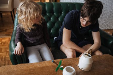 İki kardeş, ahşap bir masadaki küçük bir projeye odaklanmış yeşil bir kanepede birlikte oturuyorlar. Sakin bir öğleden sonra odasının rahat bir atmosferi vardır..