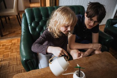 İki küçük çocuk kahve masasında küçük bir bitkiyi beslemekle meşgul. Bir çocuk bitkiyi beyaz bir sulama tenekesiyle dikkatlice sularken diğeri dikkatle izliyor..