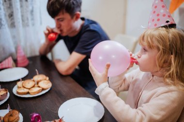 Ailesiyle birlikte bir doğum gününü kutlarken, iki kardeş masada eğleniyorlar. Kız pembe bir balon şişirirken, erkek kardeşi abur cubur yiyor..