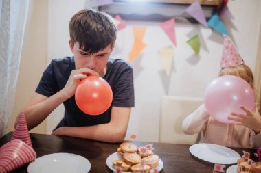 Kardeşler, renkli balonlar şişirip şenlik şapkaları giyerken tatlı ikramları paylaşarak evde neşeli bir partinin tadını çıkarıyorlar. Kahkaha ve neşe odayı doldurur birlikte kutlarken.