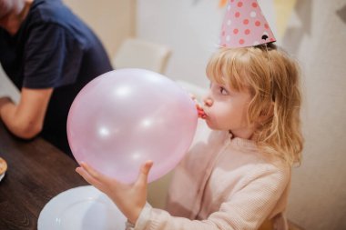 Kıvırcık saçlı bir çocuk aile toplantısında parti şapkası takarken pembe bir balonu şişiriyor. Atmosfer neşeli, kahkaha ve sıcaklıkla dolu..