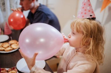 Küçük bir çocuk pembe bir balonu şişirirken diğer bir kardeş kapalı bir doğum günü partisinde eğleniyor. Renkli şapkalar ve atıştırmalıklar neşeli bir atmosfer yaratır..
