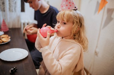 Kapalı bir ortamda, kardeşler doğum günlerini şenlikli şapkalar ve balonlarla kutlarlar. Genç bir kız pembe bir balonu şişirirken diğer kardeş yakınlarda atıştırmalık bir şeyler hazırlıyor..