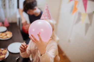 Kardeşlerin neşeli bir atmosferin tadını çıkardığı neşeli bir toplantı. Bir çocuk pembe bir balon tutarken, diğerleri de süslenmiş bir masada neşeli parti öğeleriyle atıştırmalıklar hazırlıyor..